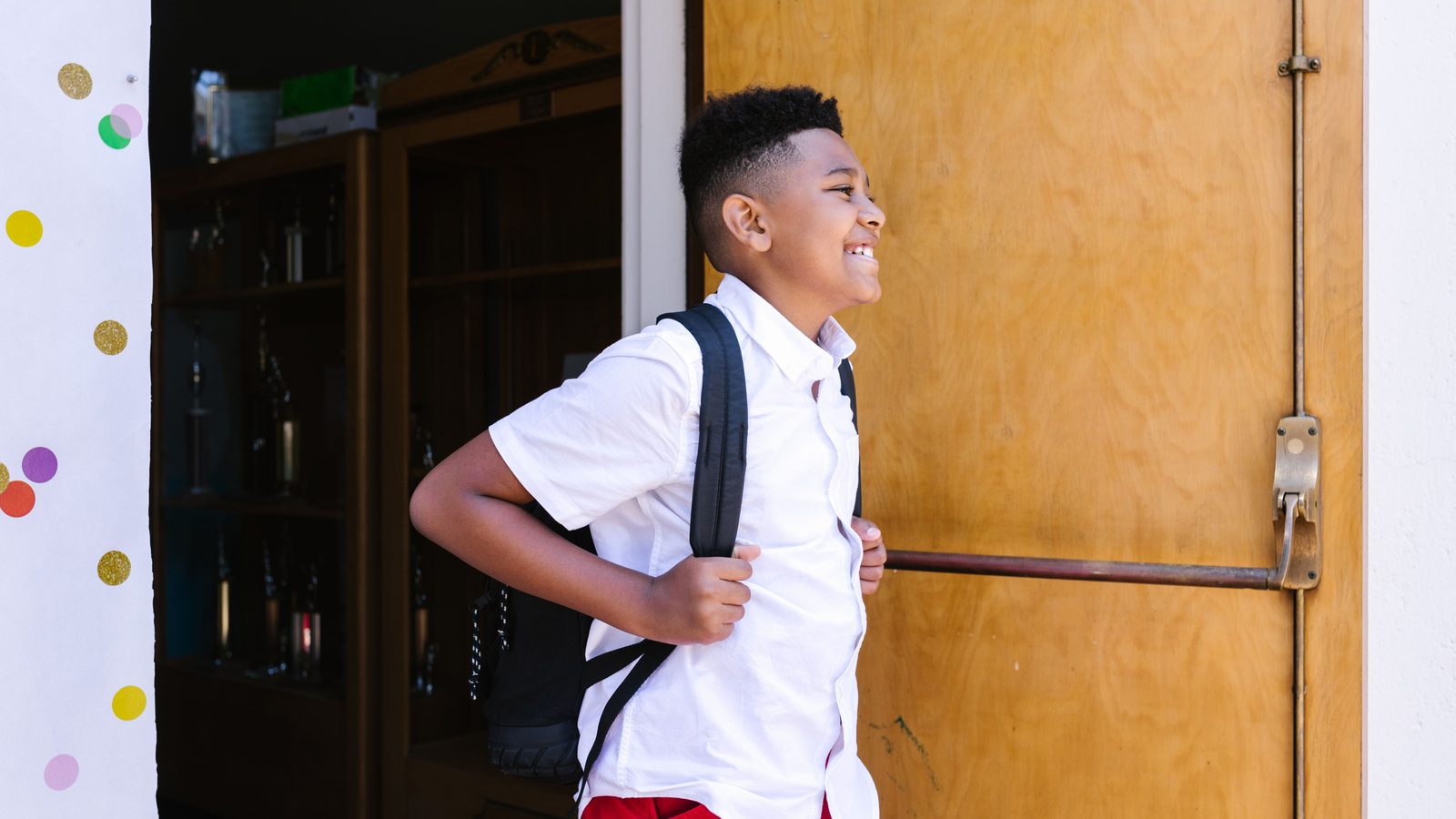 Un garçon souriant, cartable au dos, entre dans son école.