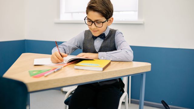 Un jeune élève à lunettes écrit à son bureau, concentré.
